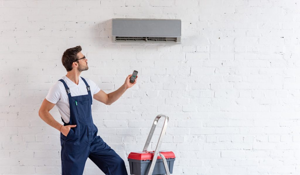 workman in overalls holding remote control while standing under minisplit AC