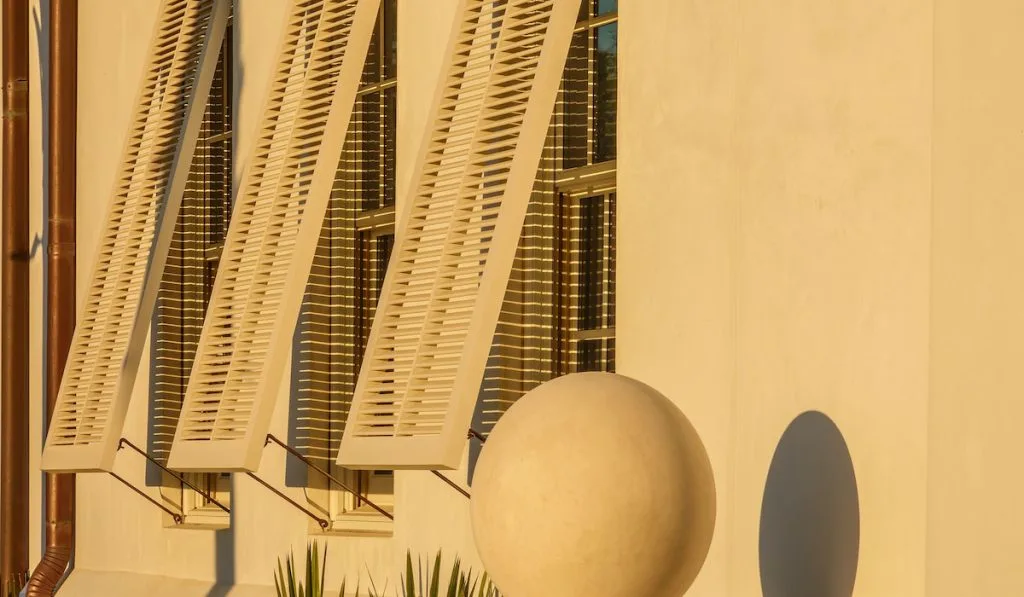 Ornamental concrete sphere, about the size of a beach ball, and its sunset shadow by three tall hurricane shutters on a concrete house 