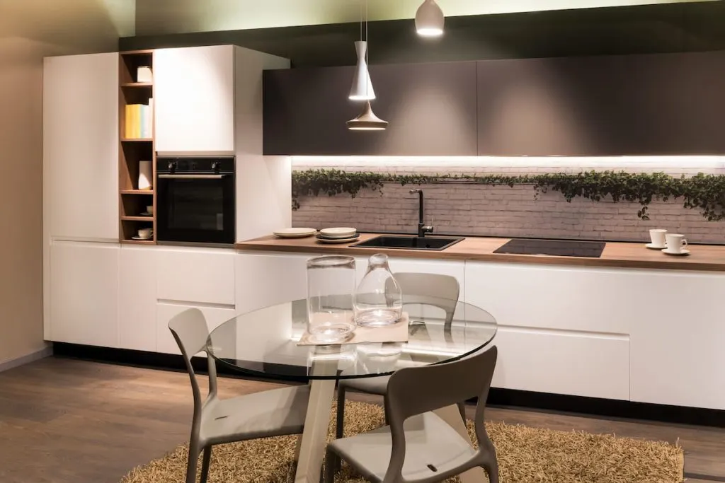 Round glass table with glassware and chairs located near cupboards and counters with built in appliances in modern kitchen at home