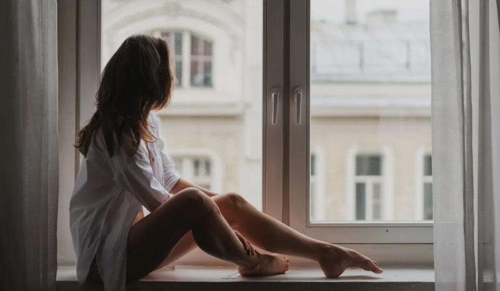  young woman with long hair in a white shirt elegantly sitting on the windowsill in an apartment