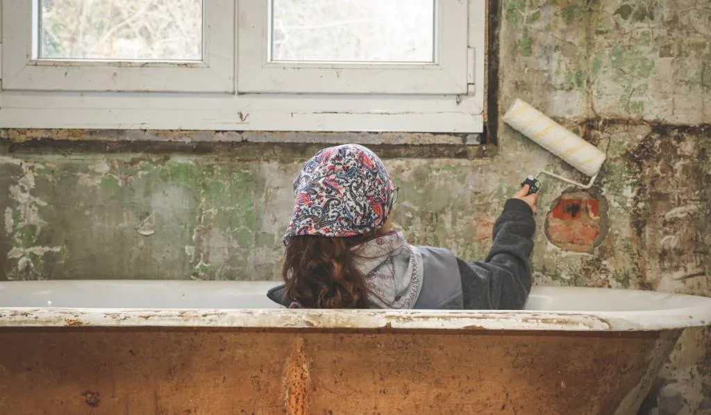 A Caucasian teenage girl in a bandana sits in the bathroom and holds a roller for for painting wall.
