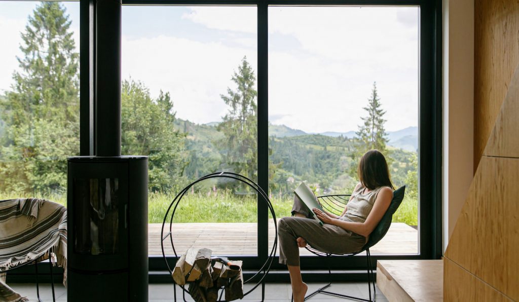 woman reading book on chair at fireplace with firewood looking through impact windows on background of mountain hills