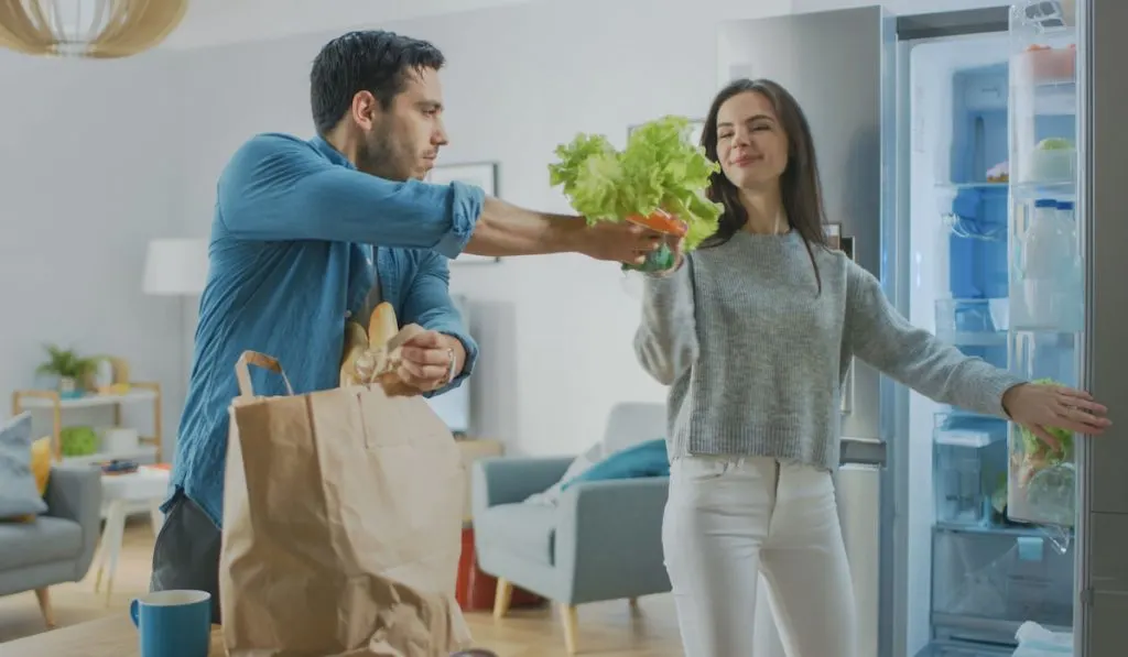 Couple stocking up the fridge, man getting groceries from paper bag and woman putting them in the fridge