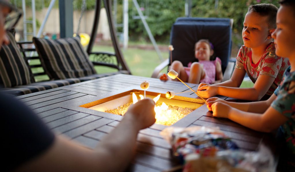 Kids sitting around a patio fire pit with flames roasting s’mores.
