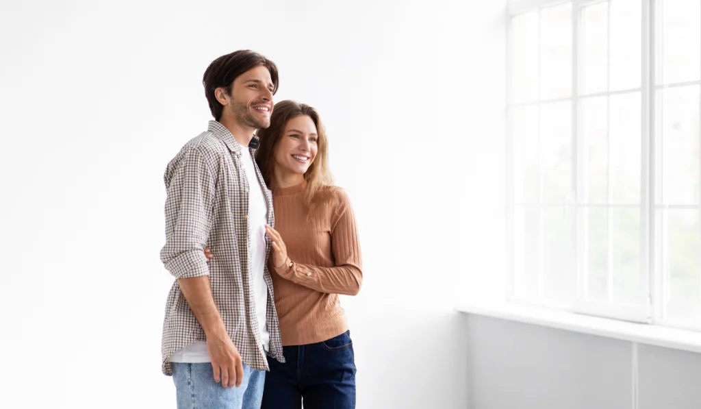 Happy glad European millennial husband and wife enjoy buying a new apartment in empty room
