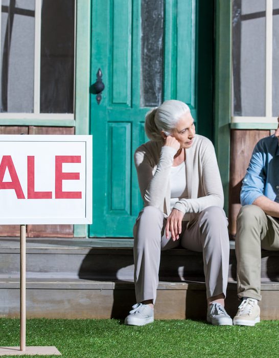 upset-senior-couple-sitting-on-porch-and-selling-their-house-house-for-sale-concept-