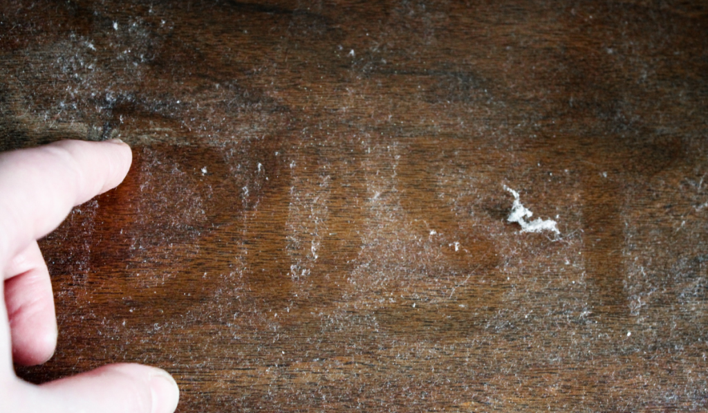 A hand writes the word "dust" on a dusty wooden surface