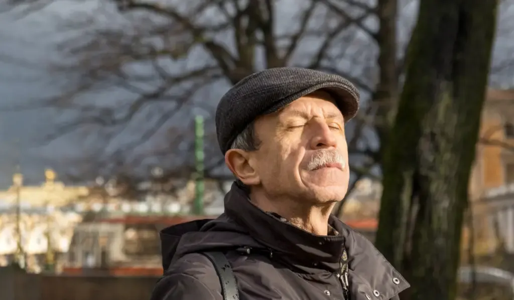 Adult man of retirement age stands in park in late autumn and exposes sun's rays