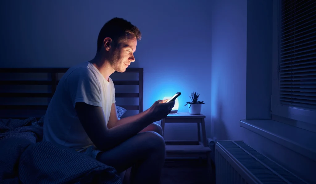 Lonely man sitting on bed in bedroom and using phone at night. Face illuminated from smartphone display.