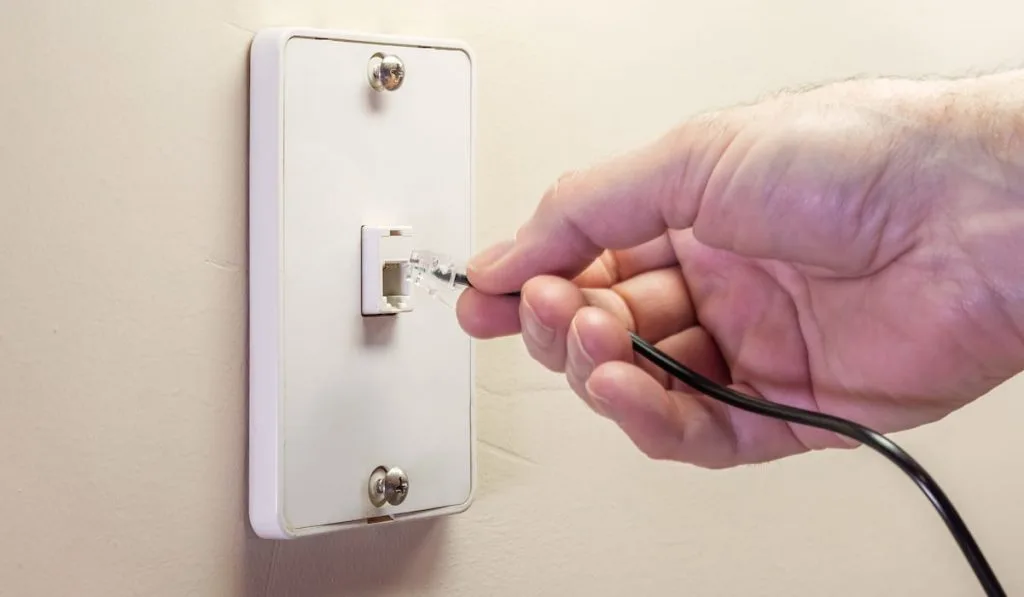 Male electrician plugging in landline telephone cord into wall mounted jack plate.