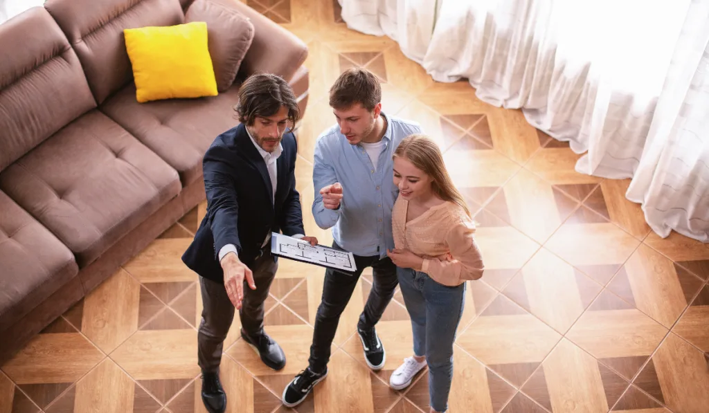 Real estate broker with house plan showing young married couple around property for sale, above view